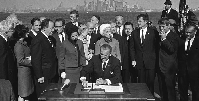 President Johnson sits in the oval office surrounded by individuals while he signs the Immigration Act of 1965 into law