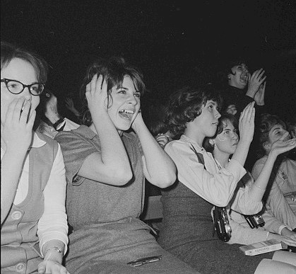 several fans clap along at a concert, one is screaming with their hands over their ears.