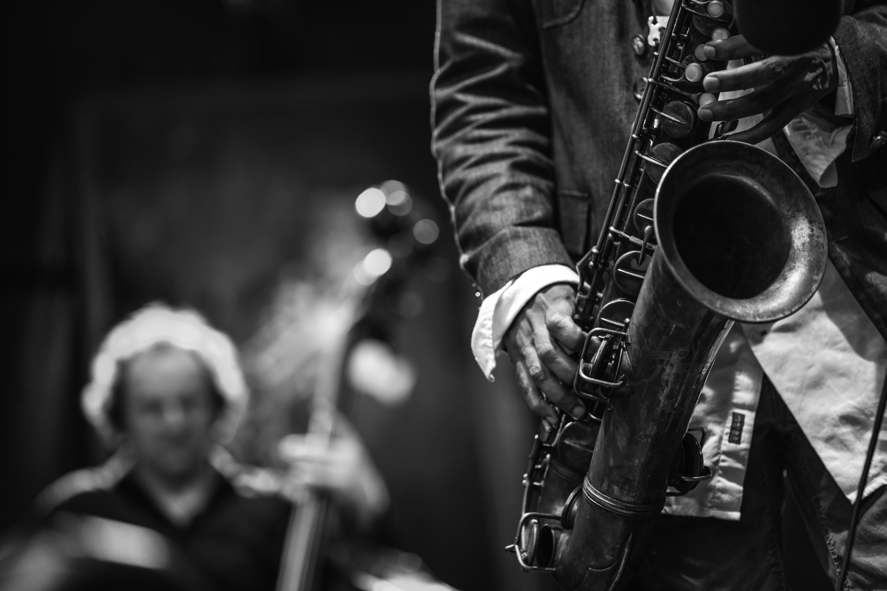 An individual plays the saxophone, all the image shows is the saxophone and an arm and hand on the keys with a string player in the background.