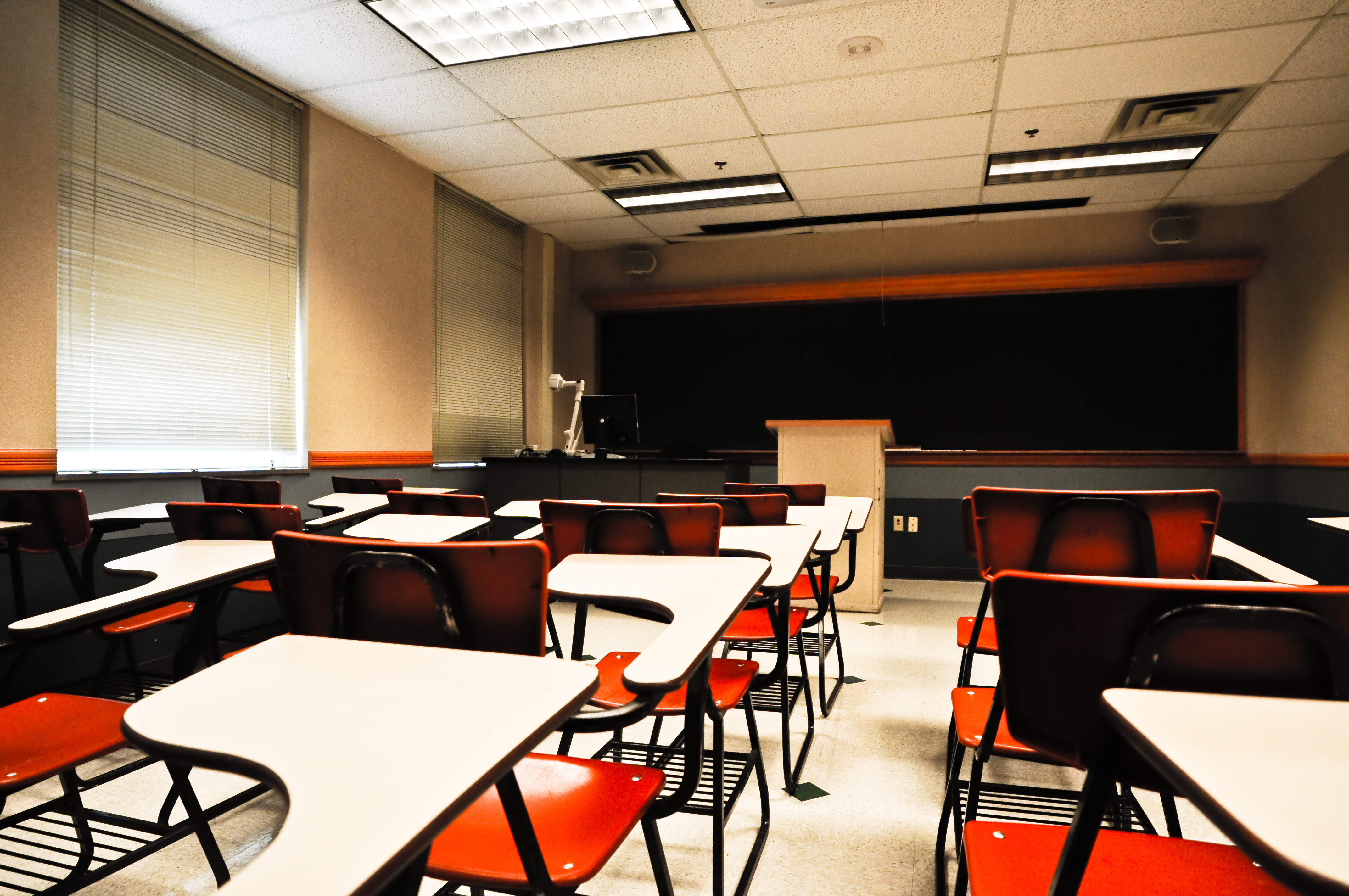An empty classroom. The image is taken from the back and shows a blackboard and podium at the front and several rows of white desks with red metal chairs for students.