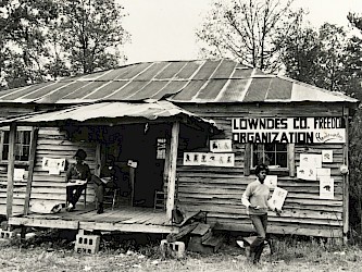 Two black individuals, one seated on the porch, one standing outside, are in front of a building that has a sign labeling it the 'Lowndes Co. Freedom House'