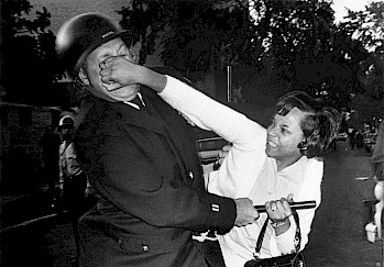 Black and white image of a black woman, in a white suit, punching a police officer in the face while holding on to her purse and the officer's baton with the same hand.