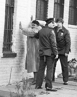 A black woman stands held against a brick wall by two white police offers. She is looking at the camera while the officers look away.