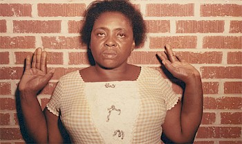 A black woman in a gingham dress stands against a brick wall with both her hands up in a defensive posture with a blank stare on her face with tears in her eyes.