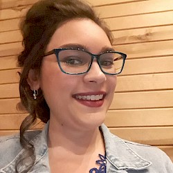 Ayoka wears teal glasses and a denim jacket and is shown smiling