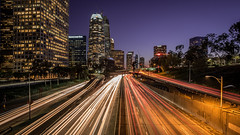 P portion of a highway with buildings behind it at dusk, with the taillights and headlights of the traffic stretched out due to time-lapse